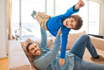 Father and son playing together on sofa in living room. Man lifting up his boy. Family staying at home due to pandemic COVID-19.